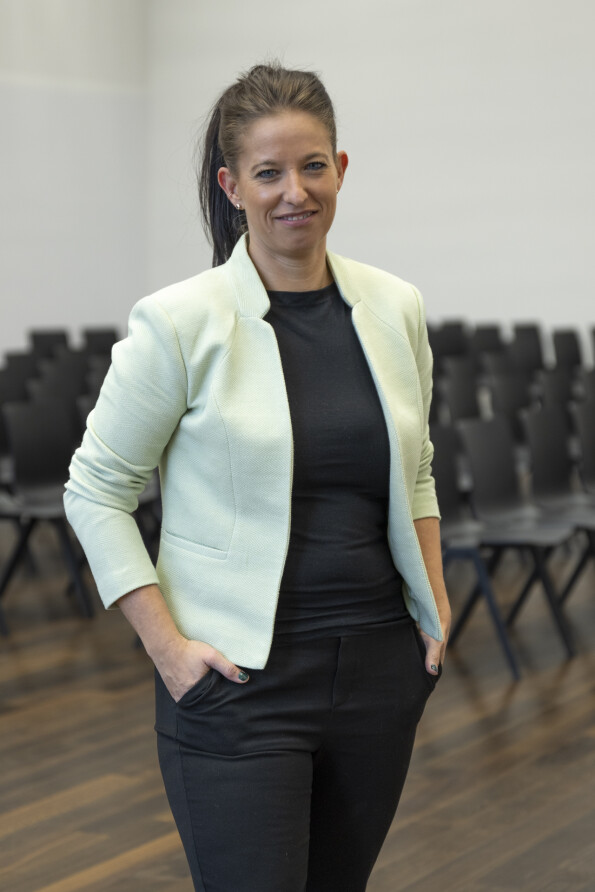 Eine Frau mit langen, dunkelbraunen Haaren und einem freundlichen Lächeln steht in einem Konferenzraum mit schwarzen Stühlen. Sie trägt ein mintgrünes Sakko, ein schwarzes Oberteil und eine schwarze Hose, mit den Händen in den Hosentaschen.